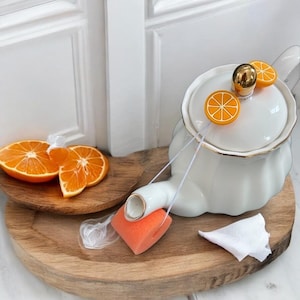 Puede incluir: Tetera blanca con un acento dorado y acentos de cítricos naranja. La tetera está en una bandeja de madera con rodajas de naranja.