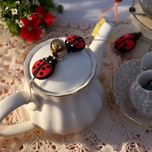 Op de afbeelding: Een witte theepot met een gouden rand en twee rode lieveheersbeestje-charms die aan de deksel zijn bevestigd.