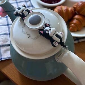 Könnte beinhalten: Eine tealfarbene Teekanne mit weißem Deckel und Griff, verziert mit zwei schwarz-weißen Bulldoggen-Anhängern. Der Deckel hat einen grauen Knopf und florale Designs. Croissants und Marmelade sind im Hintergrund.