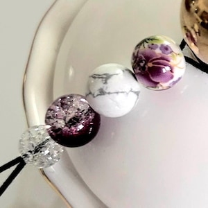 Puede incluir: Primer plano de una pulsera de cuentas con varias cuentas decorativas. Las cuentas incluyen cuentas transparentes, brillantes y blancas veteadas, así como cuentas con diseños florales morados y una cuenta morada oscura y brillante.