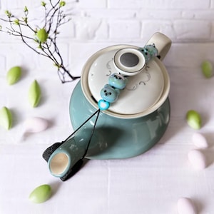 Puede incluir: Una tetera de color turquesa con tapa blanca y un cordón negro con cuentas azules y blancas unidas al pico. La tetera está sobre una superficie blanca.