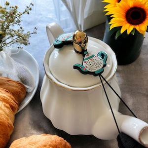 May include: White ceramic teapot with a gold trim and a black string attached to a teal blue teapot charm that says "Spilling the Tea".