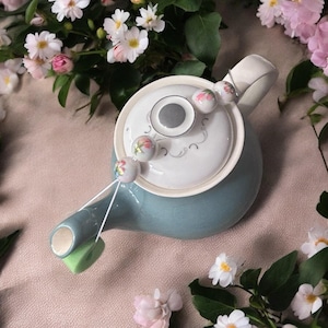 May include: A blue teapot with a white lid and floral design. A string of floral beads is attached to the lid and hangs down the side of the teapot.