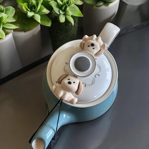 Könnte beinhalten: Eine blaue Teekanne mit einem weißen Deckel und einem schwarzen Schnürsenkelgriff. Zwei kleine braune Hundefiguren sind am Deckel befestigt.