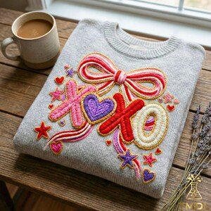 Peut inclure: Un pull gris avec un motif brodé représentant un nœud, des cœurs, des étoiles et les lettres "XOXO" en rose, rouge, violet et blanc. Le pull est posé sur une surface en bois, avec une tasse de café et des brins de lavande à proximité.