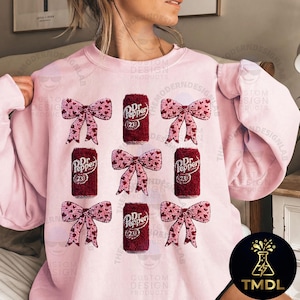 Puede incluir: Sudadera rosa claro con un diseño de latas de Dr. Pepper y lazos rosas adornados con corazones. Las latas de Dr. Pepper tienen el número 23 impreso. La sudadera es de color rosa claro.