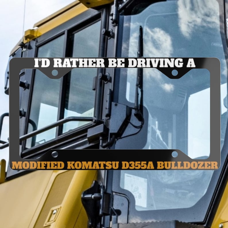 May include: Black license plate frame with the text "I'D RATHER BE DRIVING A" above "MODIFIED KOMATSU D355A BULLDOZER". The frame is mounted on a yellow and black bulldozer.