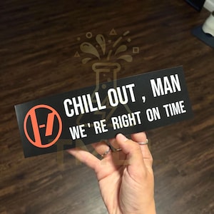 May include: A rectangular black sticker featuring the white text "CHILL OUT, MAN WE'RE RIGHT ON TIME" and an orange circular logo containing a vertical line and a dash. The design is printed on a smooth, matte surface with sharp, clean edges.