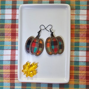 May include: A pair of wooden pumpkin earrings with a plaid pattern in shades of orange, red, green, and black. The earrings are on a white plate with a yellow maple leaf.