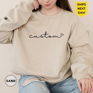 May include: A light beige sweatshirt with the word "custom" written in black cursive with a small heart symbol next to it. The sweatshirt is being worn by a person with long brown hair.