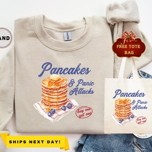 May include: A beige sweatshirt with a graphic of a stack of pancakes and the text "Pancakes & Panic Attacks" in blue. The graphic also includes blueberries and a butter pat. The text "Buy one get one" is also included in red.