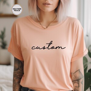以下が含まれることがあります： ピーチ色のTシャツに、黒い筆記体で「custom」と書かれています。