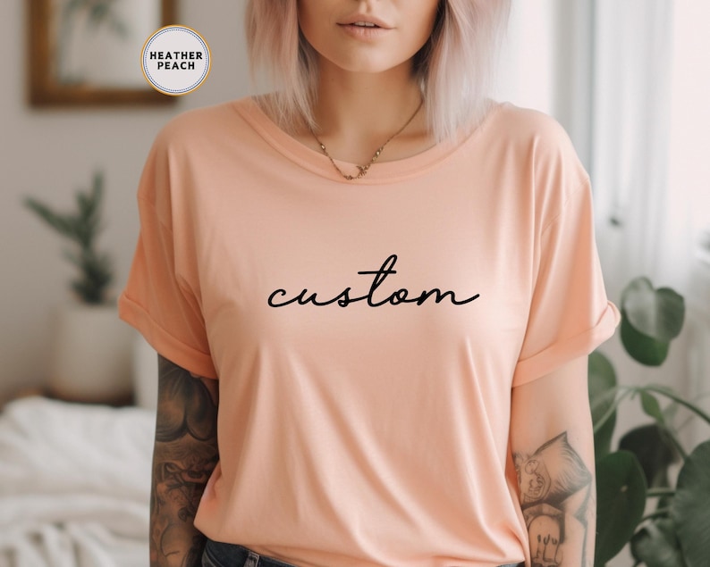以下が含まれることがあります： ピーチ色のTシャツに、黒い筆記体で「custom」と書かれています。
