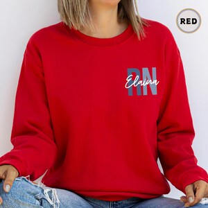May include: Red sweatshirt with the text "RN Elaina" in grey and blue lettering.