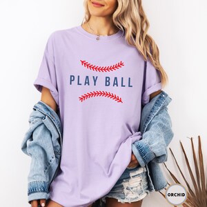 May include: A lavender t-shirt with a baseball graphic and the text "PLAY BALL" in blue lettering.