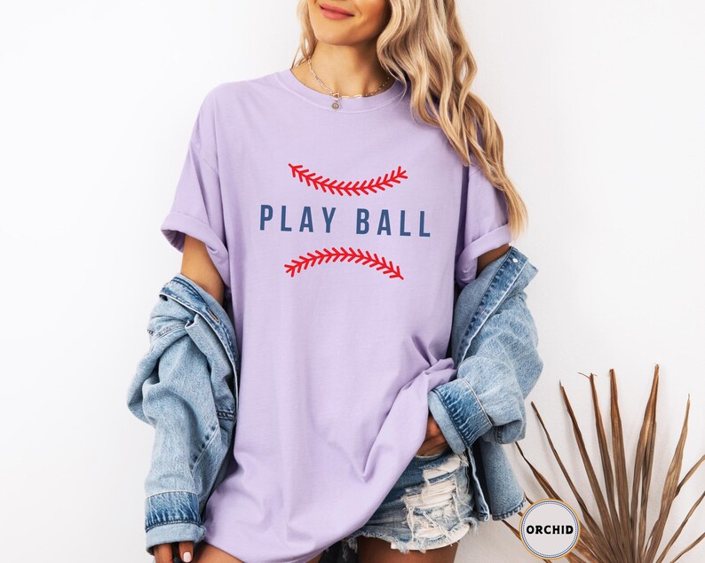 May include: A lavender t-shirt with a baseball graphic and the text "PLAY BALL" in blue lettering.