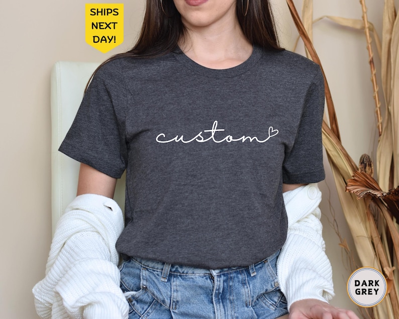 以下が含まれることがあります： 「custom」という単語が白い筆記体で書かれた、濃いグレーのTシャツ。単語の後に小さなハートマークがついています。