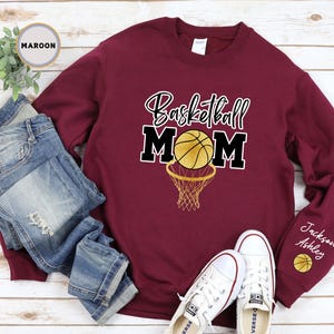 May include: Maroon sweatshirt with the text "Basketball Mom" in gold and black lettering. A gold basketball with black lines is inside a black basketball hoop. The sweatshirt has a white tag on the collar and the text "Jackson Ashley" on the sleeve with a small basketball graphic.