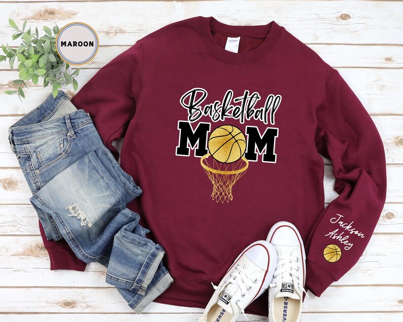 May include: Maroon sweatshirt with the text "Basketball Mom" in gold and black lettering. A gold basketball with black lines is inside a black basketball hoop. The sweatshirt has a white tag on the collar and the text "Jackson Ashley" on the sleeve with a small basketball graphic.