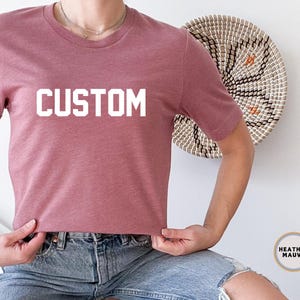 以下が含まれることがあります： 前面に白い文字で「CUSTOM」がプリントされた、ヘザーモーヴのTシャツです。