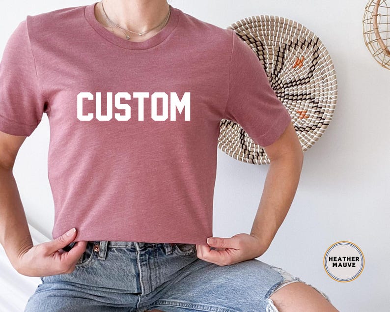 以下が含まれることがあります： 前面に白い文字で「CUSTOM」がプリントされた、ヘザーモーヴのTシャツです。