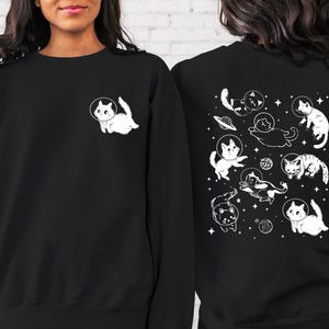 Peut inclure: Sweat-shirt noir avec un motif blanc de chats dans l'espace au dos et un seul chat dans l'espace sur le devant. Les chats portent des casques d'astronautes.