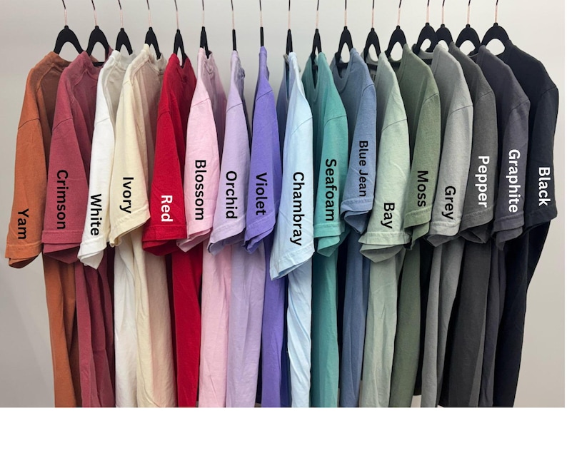 May include: A row of 15 t-shirts hanging on a rack, showcasing a variety of colors, including crimson, white, ivory, red, blossom, orchid, violet, chambray, seafoam, blue jean, bay, moss, grey, pepper, graphite, and black.