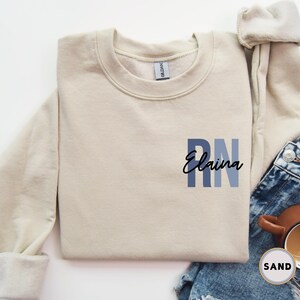May include: A beige sweatshirt with the text "RN Elaina" in blue and black lettering.