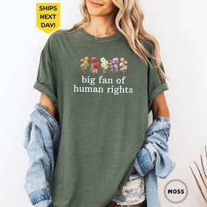 May include: Olive green t-shirt with the text "big fan of human rights" in white. The shirt features a floral design above the text. A denim jacket is draped over the arms. The shirt is a moss color.