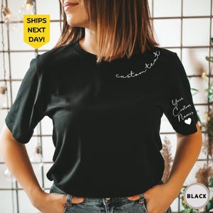 May include: Black short-sleeved t-shirt with custom text on the back and sleeve. The sleeve features the text "Your Custom Names" and a white heart. The shirt has a round neckline.