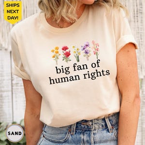 May include: Cream-colored t-shirt with the text "big fan of human rights" in black. Above the text are watercolor illustrations of yellow, red, white, and purple flowers. The shirt is worn with blue jeans.