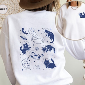 Peut inclure: Sweat-shirt blanc avec un imprim&eacute; bleu de chats portant des casques d'astronautes et flottant dans l'espace. Le texte "WHITE" est visible dans le coin sup&eacute;rieur gauche de l'image.
