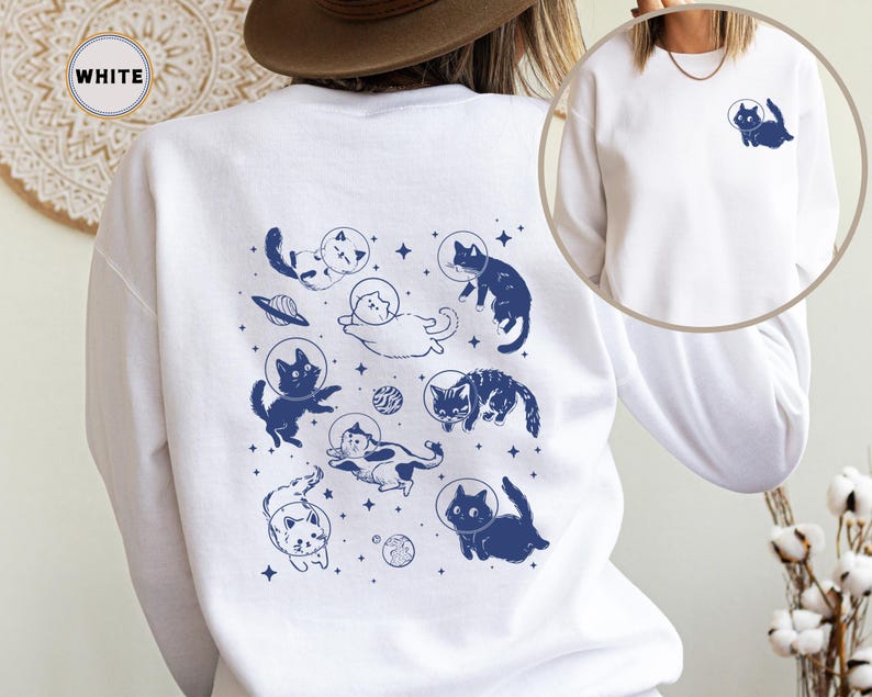 Peut inclure: Sweat-shirt blanc avec un imprim&eacute; bleu de chats portant des casques d'astronautes et flottant dans l'espace. Le texte "WHITE" est visible dans le coin sup&eacute;rieur gauche de l'image.