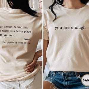 May include: A beige t-shirt with the text "dear person behind me, the world is a better place with you in it. love, the person in front of you" on the back and "you are enough." on the front.
