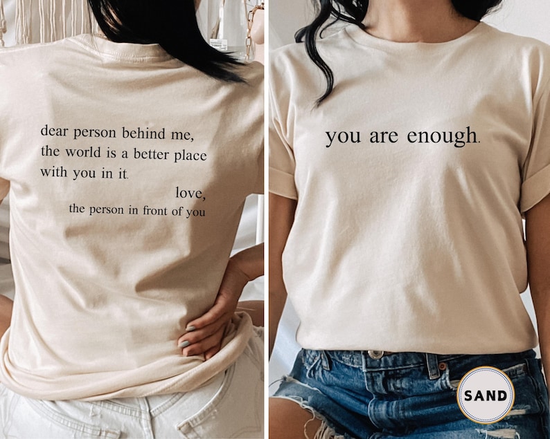 May include: A beige t-shirt with the text "dear person behind me, the world is a better place with you in it. love, the person in front of you" on the back and "you are enough." on the front.