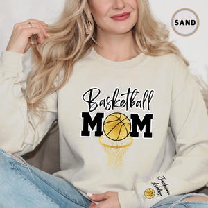 May include: A light beige sweatshirt with the text "Basketball Mom" in black with a gold basketball and net graphic. The sweatshirt has a gold embroidered name tag on the sleeve that says "Jackson Ashley".