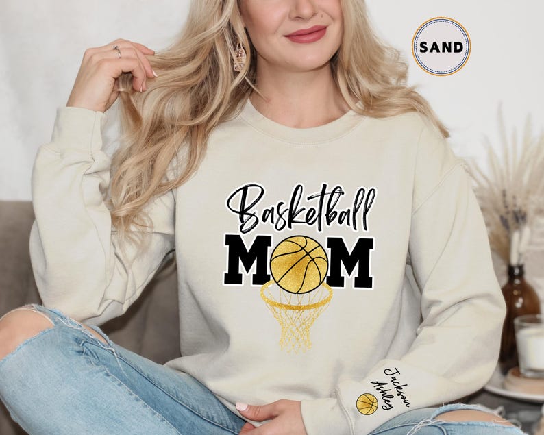 May include: A light beige sweatshirt with the text "Basketball Mom" in black with a gold basketball and net graphic. The sweatshirt has a gold embroidered name tag on the sleeve that says "Jackson Ashley".