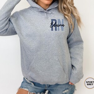 May include: A grey hooded sweatshirt with the text "RN Elaina" printed on the front. The sweatshirt is a popular style for nurses.