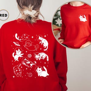 Peut inclure: Sweat-shirt rouge avec un motif blanc de chats dans l'espace. Le motif repr&eacute;sente des chats portant des casques d'astronautes et flottant dans l'espace avec des plan&egrave;tes et des &eacute;toiles. Le texte "RED" est visible dans le coin sup&eacute;rieur gauche de l'image.