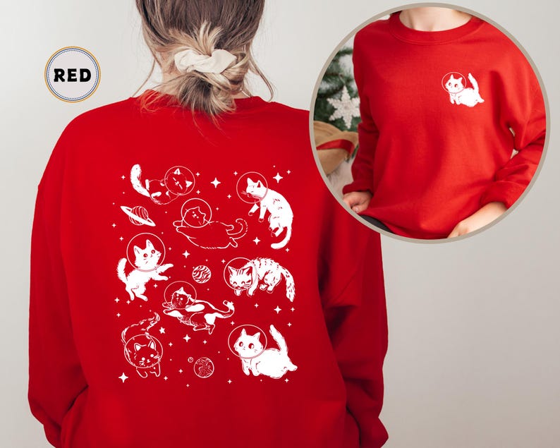 Peut inclure: Sweat-shirt rouge avec un motif blanc de chats dans l'espace. Le motif repr&eacute;sente des chats portant des casques d'astronautes et flottant dans l'espace avec des plan&egrave;tes et des &eacute;toiles. Le texte "RED" est visible dans le coin sup&eacute;rieur gauche de l'image.
