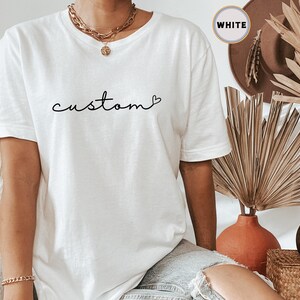 以下が含まれることがあります： 「custom」という単語が黒色の筆記体で書かれた白いTシャツ。単語の後に小さなハートマークが描かれている。