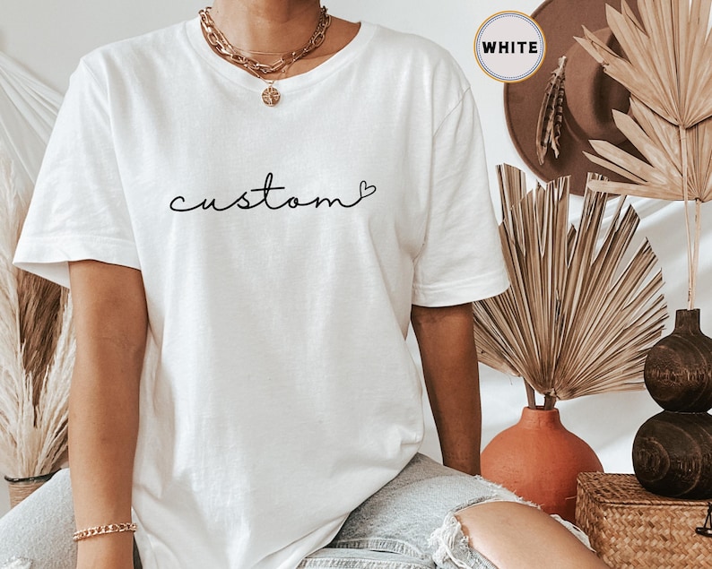 以下が含まれることがあります： 「custom」という単語が黒色の筆記体で書かれた白いTシャツ。単語の後に小さなハートマークが描かれている。