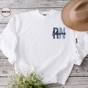 May include: White long-sleeved sweatshirt with a blue and grey ombre design that says "RN Elaina" on the front.