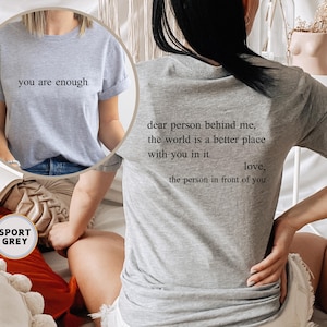 May include: Two gray t-shirts with different text printed on them. The first shirt says "you are enough." The second shirt says "dear person behind me, the world is a better place with you in it love, the person in front of you".