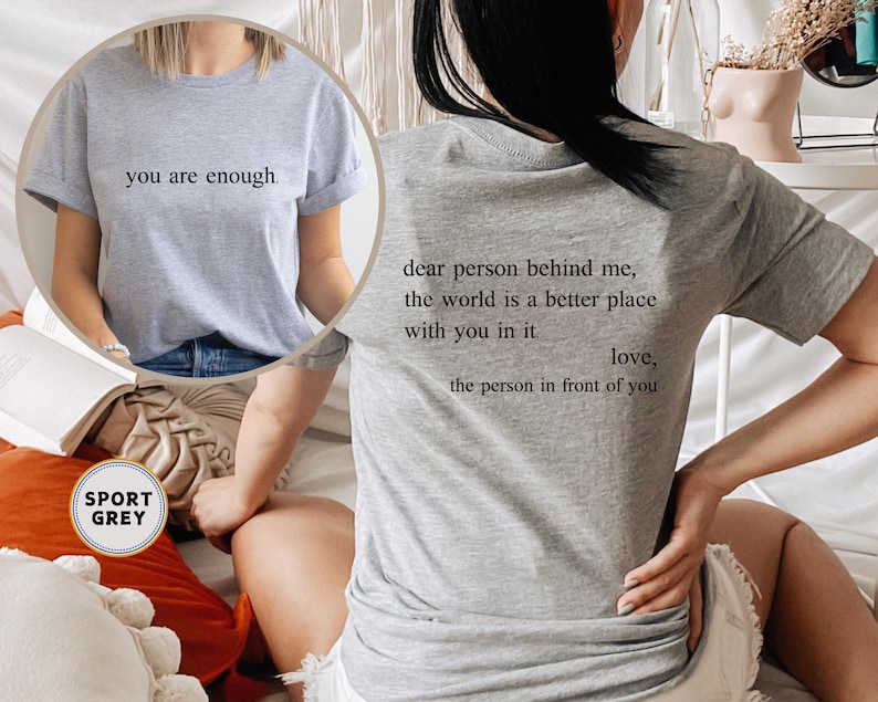 May include: Two gray t-shirts with different text printed on them. The first shirt says "you are enough." The second shirt says "dear person behind me, the world is a better place with you in it love, the person in front of you".