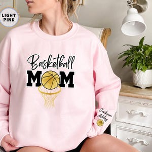 May include: Light pink sweatshirt with the text "Basketball Mom" in black with a gold basketball and hoop graphic. The sweatshirt has the names "Jackson" and "Ashley" written on the sleeve.