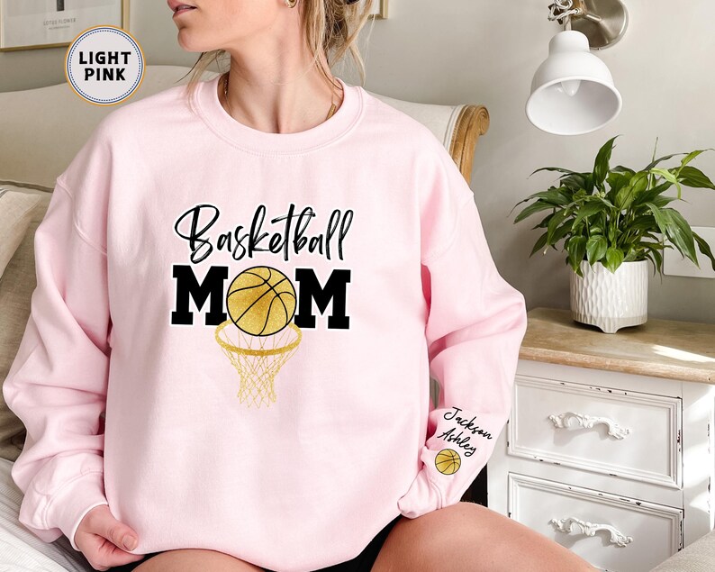 May include: Light pink sweatshirt with the text "Basketball Mom" in black with a gold basketball and hoop graphic. The sweatshirt has the names "Jackson" and "Ashley" written on the sleeve.