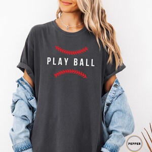 May include: A dark gray t-shirt with the text "PLAY BALL" in white letters. The text is surrounded by a red baseball stitch design.