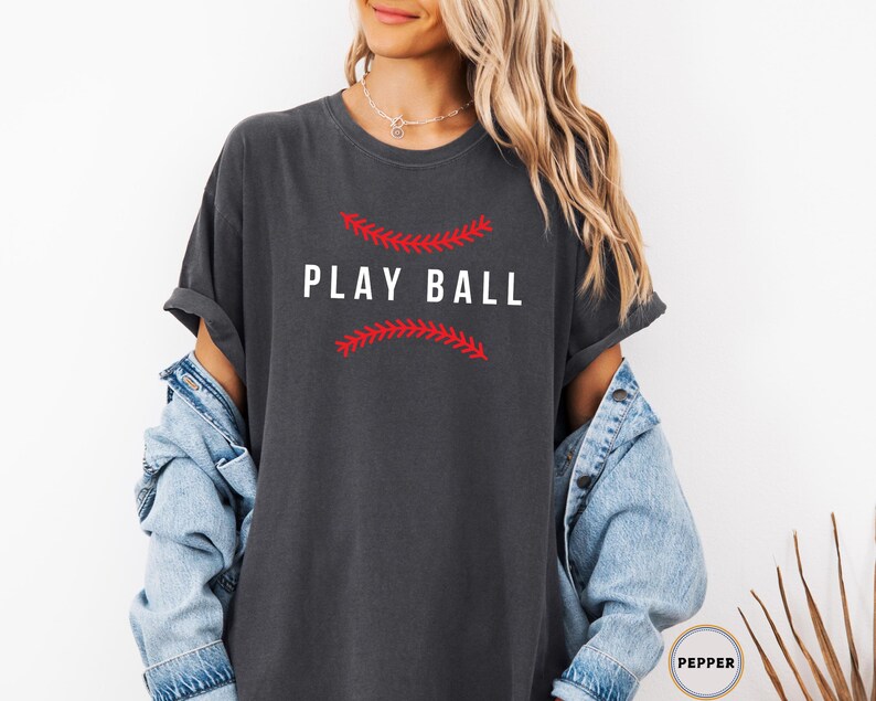 May include: A dark gray t-shirt with the text "PLAY BALL" in white letters. The text is surrounded by a red baseball stitch design.