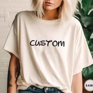 以下が含まれることがあります： 前面に黒い文字で「CUSTOM」とプリントされた白いTシャツです。